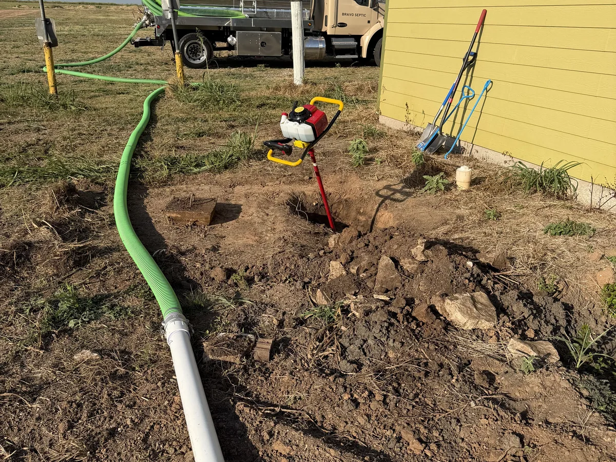 A septic tank access uncovered in a yard with digging tools and the pump truck behind